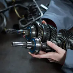 Dłonie trzymające wałek z zębatkami i łożyskami, na tle innych części mechanicznych.