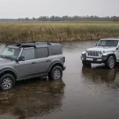 Szary Ford Bronco i biały Jeep Wrangler stoją w wodzie na tle łąki.