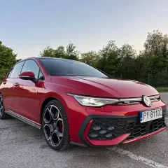 Czerwony Volkswagen Golf GTI na parkingu, z zielenią w tle.