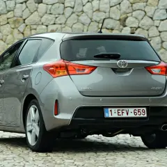 Tył szarego samochodu Toyota Auris z belgijską tablicą rejestracyjną na tle kamiennej ściany.