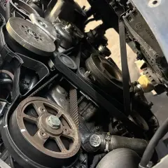 Widok z bliska na mechanizm napędu paska w samochodzie, z widocznymi kołami pasowymi i paskami.