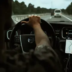 Widok z wnętrza samochodu: ręka na kierownicy, nawigacja na telefonie, droga przed samochodem.