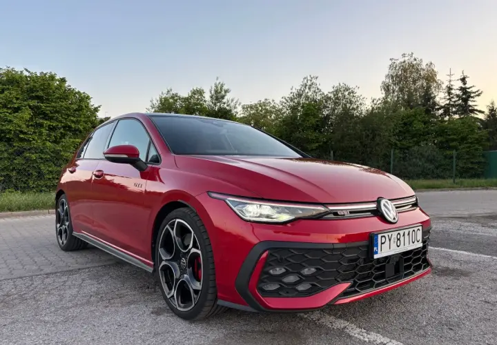 Czerwony Volkswagen Golf GTI na parkingu, z zielenią w tle.