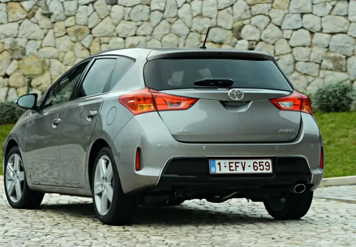 Tył szarego samochodu Toyota Auris z belgijską tablicą rejestracyjną na tle kamiennej ściany.