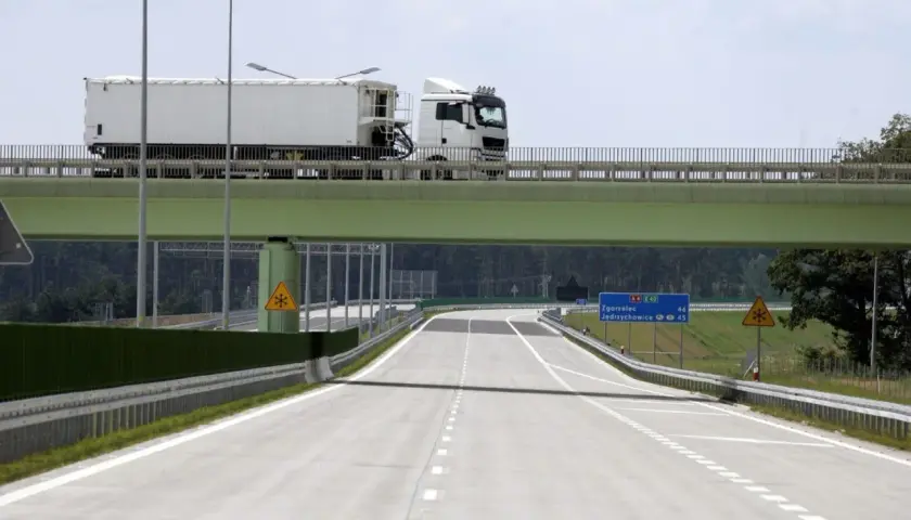 Ciężarówka na zielonym wiadukcie nad pustą autostradą z tablicami kierunkowymi.