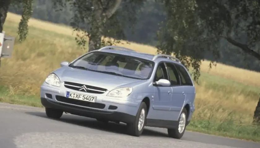 Jasnoniebieskie kombi Citroen na zakręcie drogi, z drzewami i polem w tle.