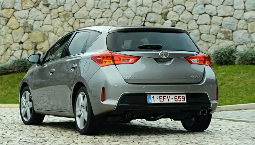 Tył szarego samochodu Toyota Auris z belgijską tablicą rejestracyjną na tle kamiennej ściany.